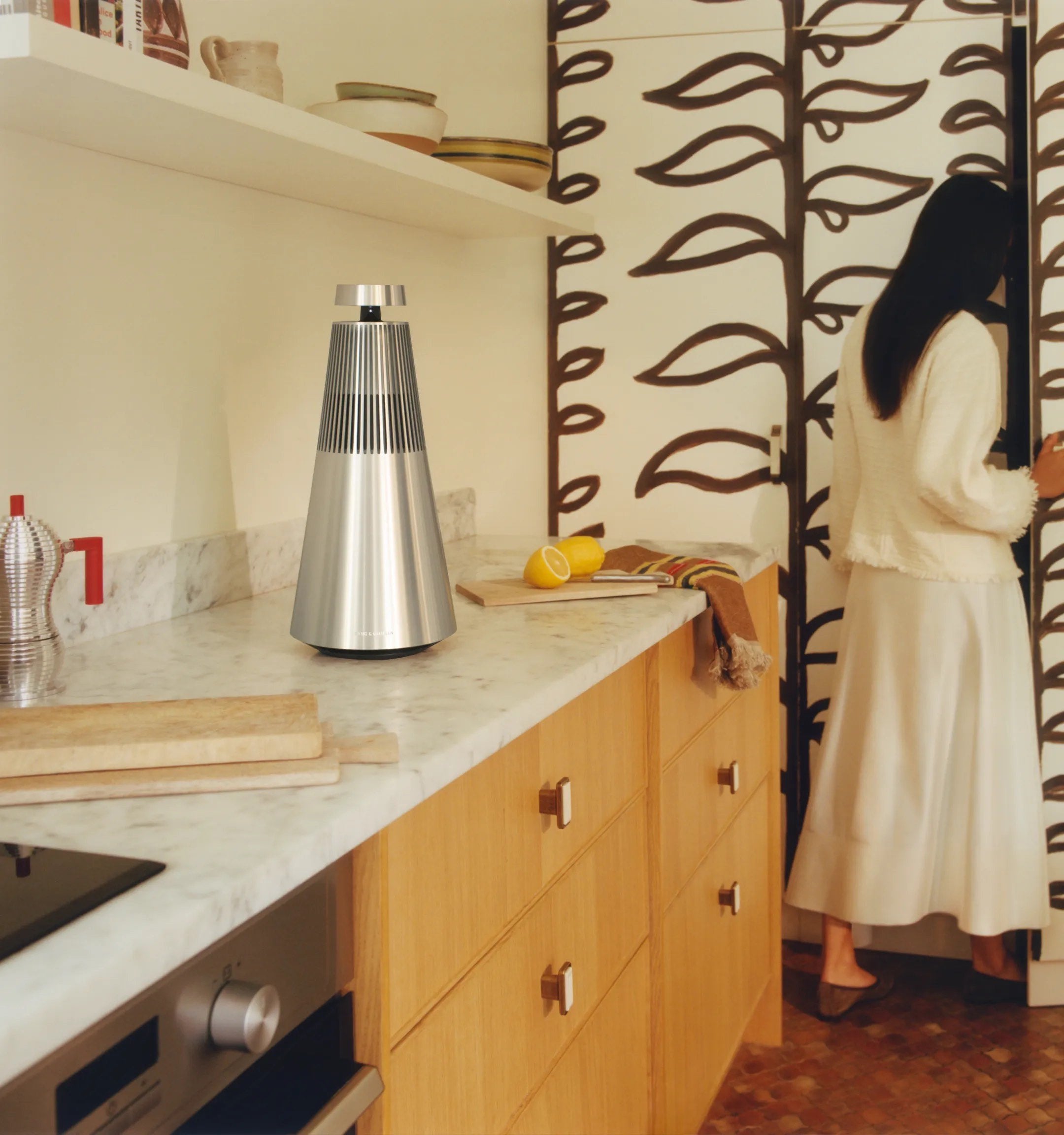Beosound 2 din aluminiu natural pe un blat de marmură, într-o bucătărie cu mobilier de stejar și tapet pictat.