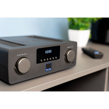 SVS Prime Wireless Pro Soundbase, aparatul pe o masă de lemn, cu o boxă pasivă în fundal și telecomanda alături