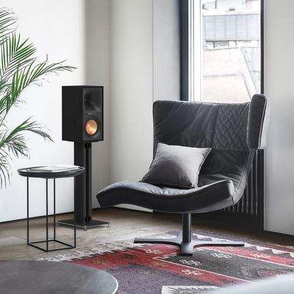 Boxa Klipsch R-50M fara grila, pe un stativ negru langa un fotoliu, intr-o camera luminoasa