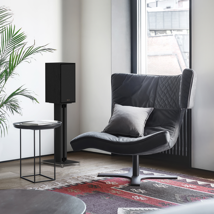 Boxa Klipsch R-50M cu grila montata, pe un stativ negru langa un fotoliu