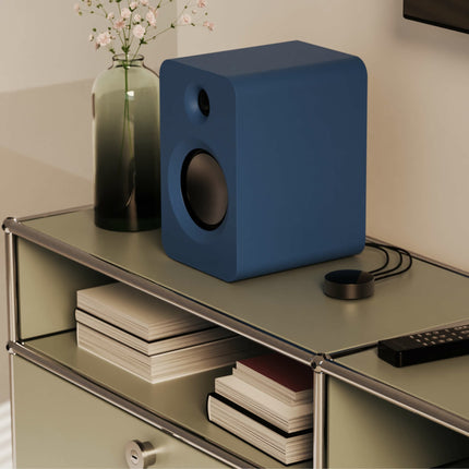 Boxă Kanto Audio REN albastră pe o comodă, lângă telecomandă și cărți