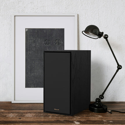 Boxa activa Klipsch R-50PM in finisaj negru, cu grila montata, pe o masa de lemn langa un tablou si o lampa de birou