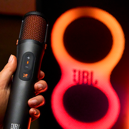 Microfon JBL EasySing Mics ținut în mână, în fața luminilor unei boxe JBL PartyBox