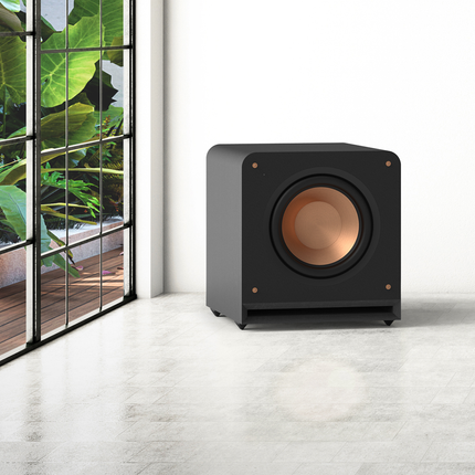 Subwooferul Klipsch RP-1200SW, fără grilă, lângă o fereastră mare, cu difuzorul Cerametallic aramiu vizibil